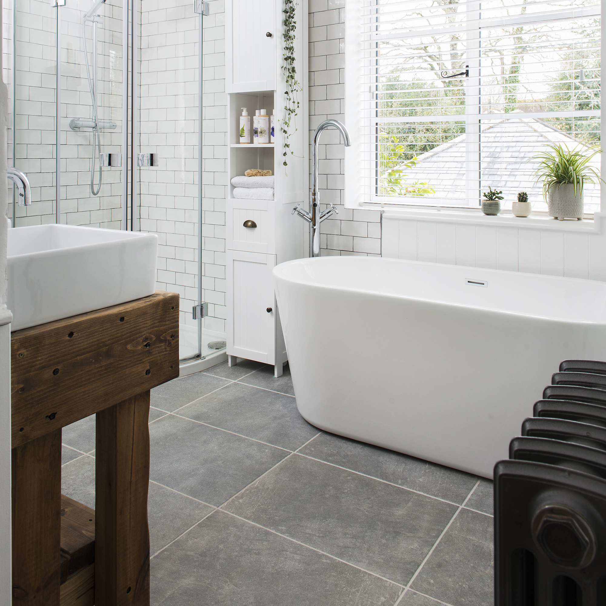 Cuarto de baño con paredes, bañera, lavabo y armarios blancos, ducha de cristal, suelo de piedra gris, radiador y luz negros y base de lavabo de madera